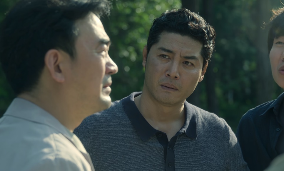 O detetive Dong-cheol olhando de lado, com outro homem em primeiro plano, ao ar livre