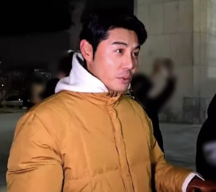 O ator Lee Kwan-hoon de jaqueta amarela e moletom branco com capuz, à noite, na rua