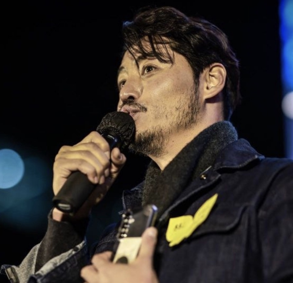 O ator Lee Kwan-hoon, de barba, falando ao microfone em um palco à noite