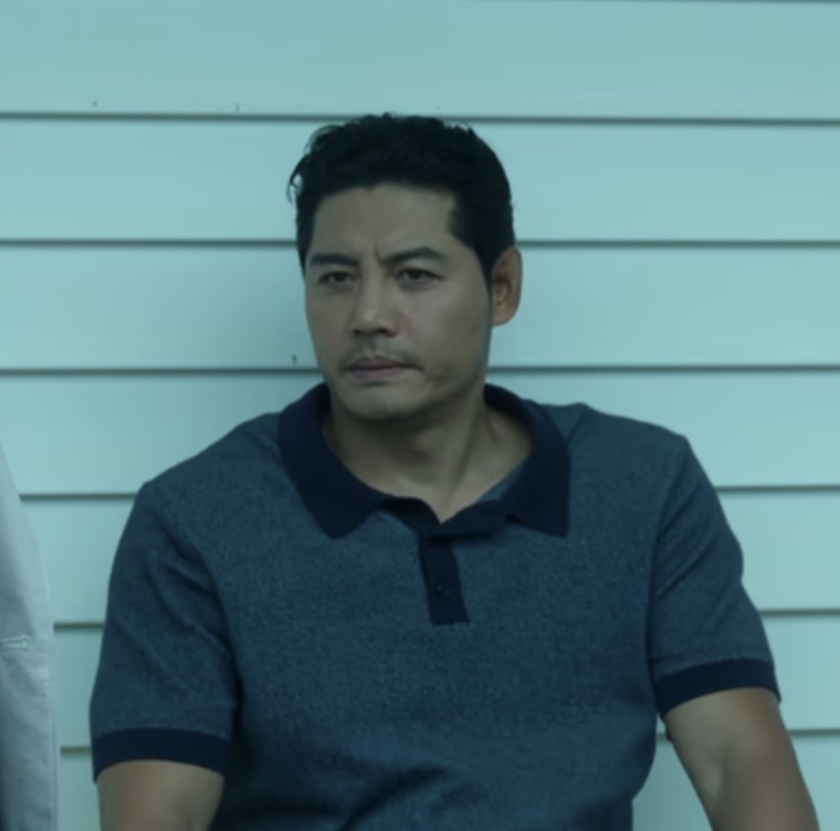 Close do detetive Dong-cheol, de camisa polo azul, encostado em uma parede clara