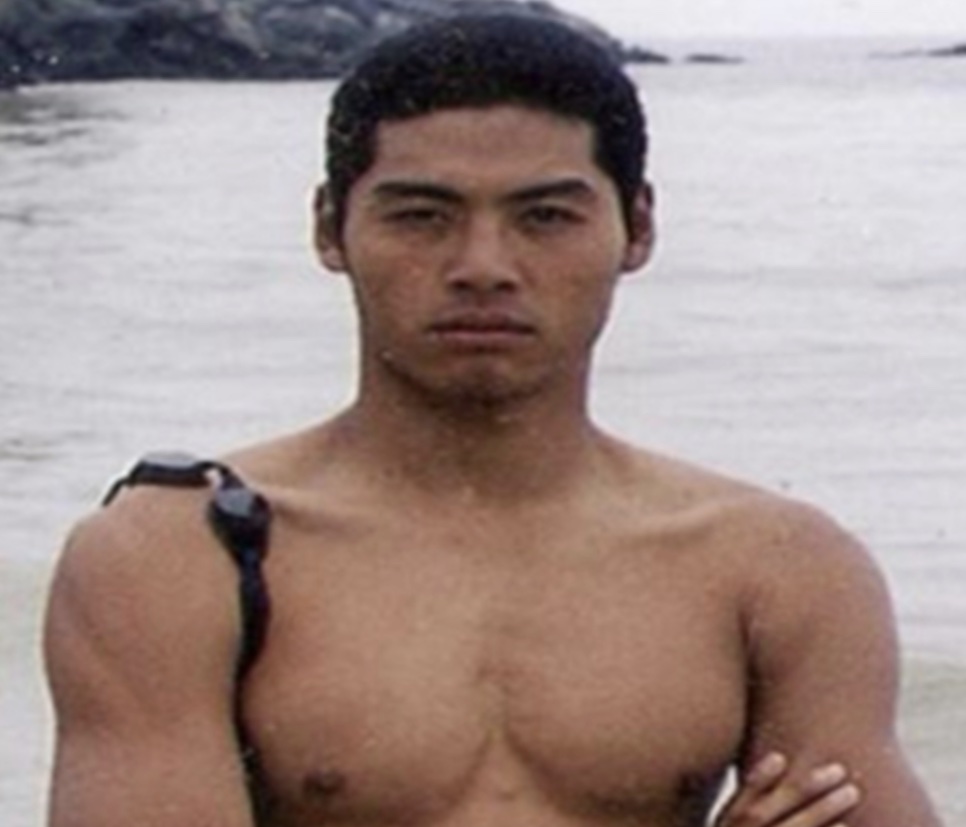 Foto antiga do ator Lee Kwan-hoon, bem mais magro, sem camisa e de braços cruzados na beira do mar
