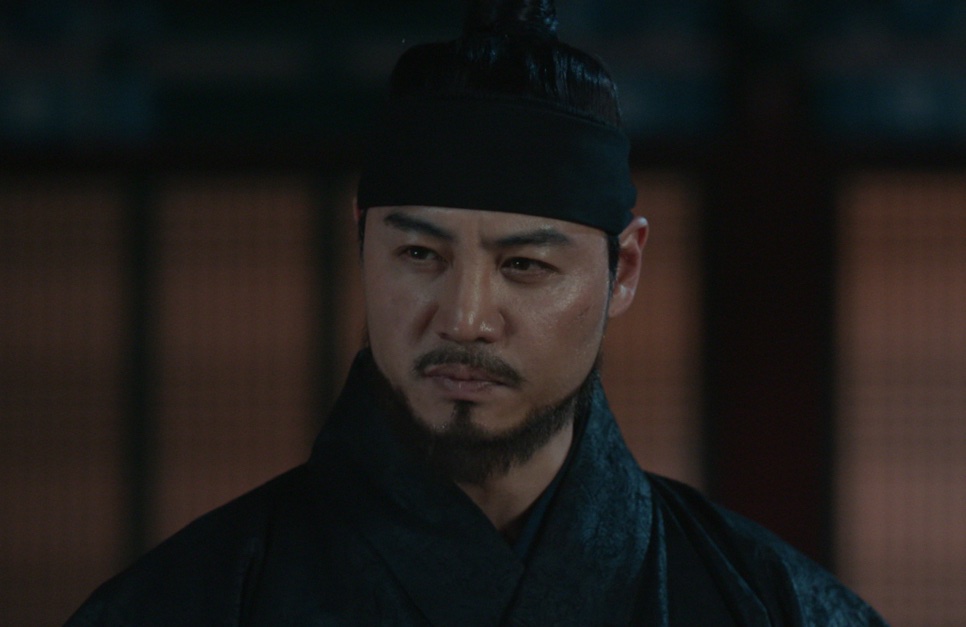 O ator Lee Kwan-hoon de chapéu preto e roupa de época, com expressão séria, no papel de Deok-chul em Bon Appétit, Vossa Majestade