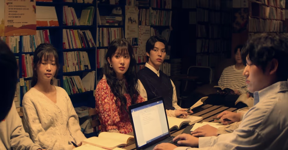 Integrantes do clube de literatura reunidos em volta de uma mesa, entre estantes de livros, com a Min-yeong ao centro