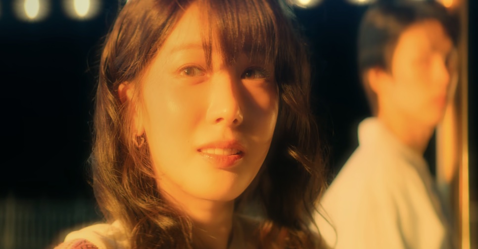 So Min-yeong sob uma luz dourada, com um homem desfocado ao fundo