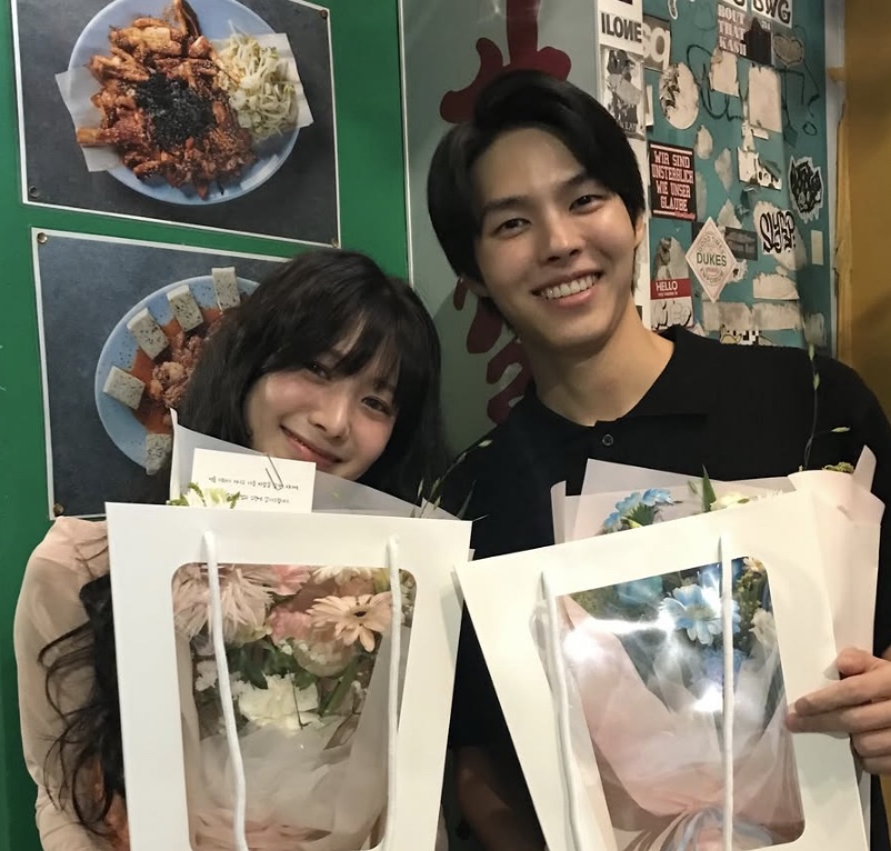 Song Su-yi e um ator do elenco segurando buquês de flores, sorrindo