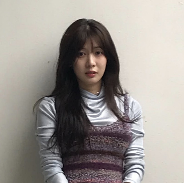 Song Su-yi com o figurino da So Min-yeong, blusa de gola alta e colete de tricô, encostada numa parede