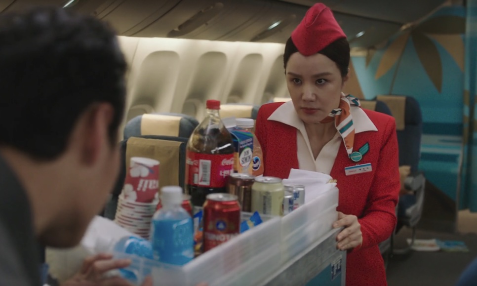 A Mi-young, vestida de comissária, empurrando o carrinho de bebidas no corredor do avião