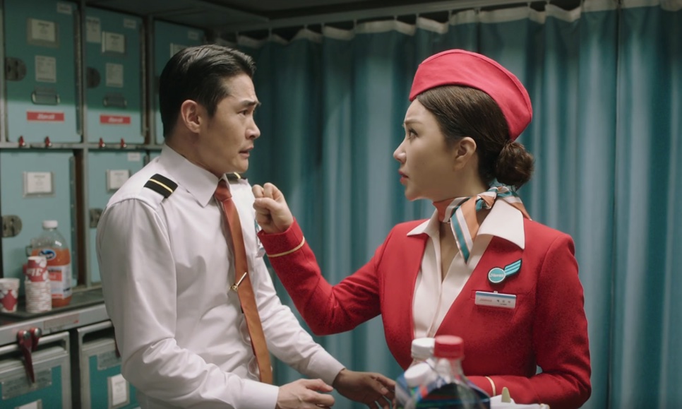 A Mi-young, de uniforme vermelho de comissária, frente a frente com um tripulante de camisa branca na área de serviço do avião