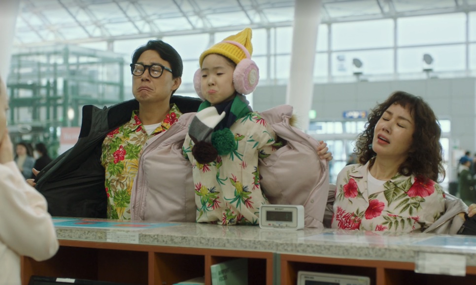 Seok-hwan, a filha Na-ri e a Mi-young, de camisas floridas e casacos de inverno, no balcão do aeroporto