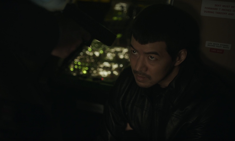 Ri Cheol-seung (Lee Sang-yoon) na cabine escura do avião, com as luzes da cidade lá embaixo pela janela