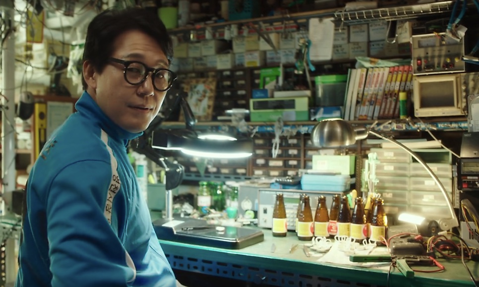 Park Sung-woong de óculos e agasalho azul, no papel do Seok-hwan, na loja de conserto de computadores cheia de peças