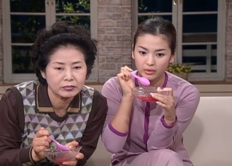 Song Hye-kyo jovem, de pijama lilás, comendo uma sobremesa ao lado de uma senhora