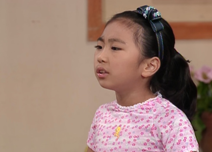 Mi-dal-i, menina de laço no cabelo e blusa florida rosa, falando com expressão brava