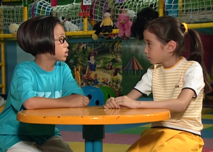 Su-ji sentada numa mesinha amarela conversando com um menino de óculos