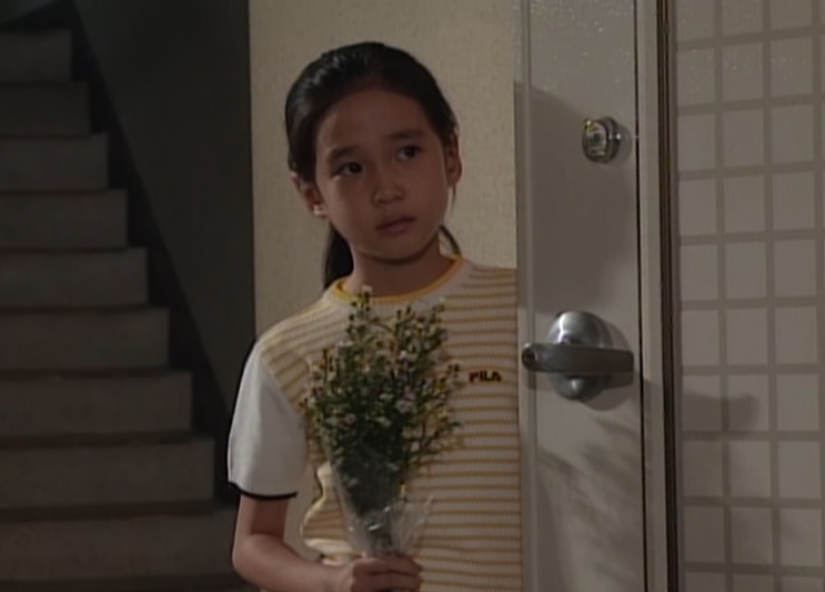 Su-ji parada na porta de um apartamento, segurando flores