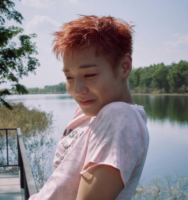 Park Ji-hoon de cabelo vermelho e camisa clara, à beira de um lago