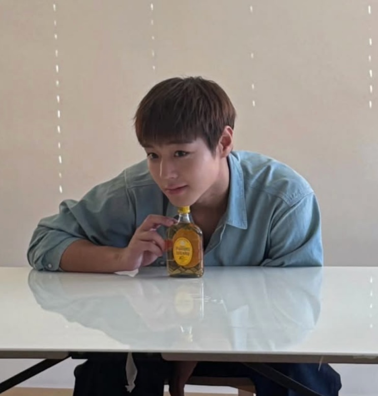 Park Ji-hoon de camisa azul-clara sentado a uma mesa, com uma garrafa à frente
