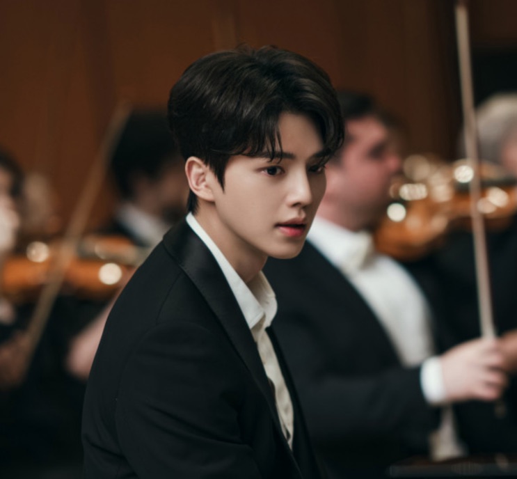 Song Kang de terno escuro no meio de uma orquestra, olhando para o lado