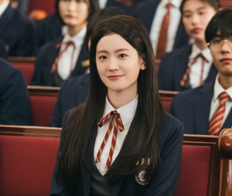 Jang Gyu-ri sentada na plateia de um auditório, entre outros alunos de uniforme