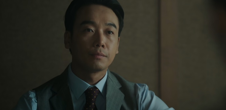 Jin Seong-jun de terno escuro, sentado e olhando para o lado