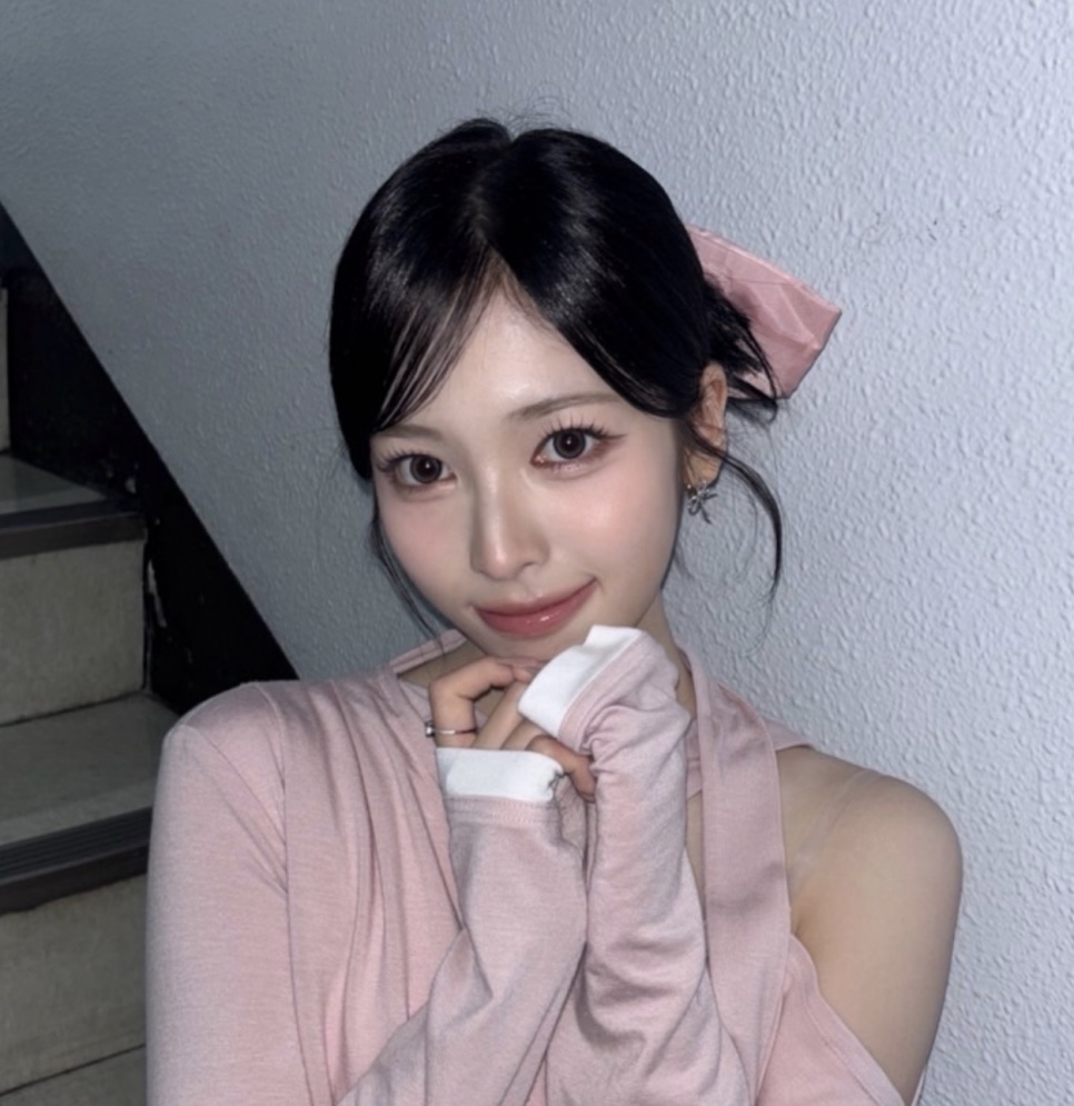 May, do Rescene, de cabelo escuro preso com um laço rosa, apoiando o queixo na mão