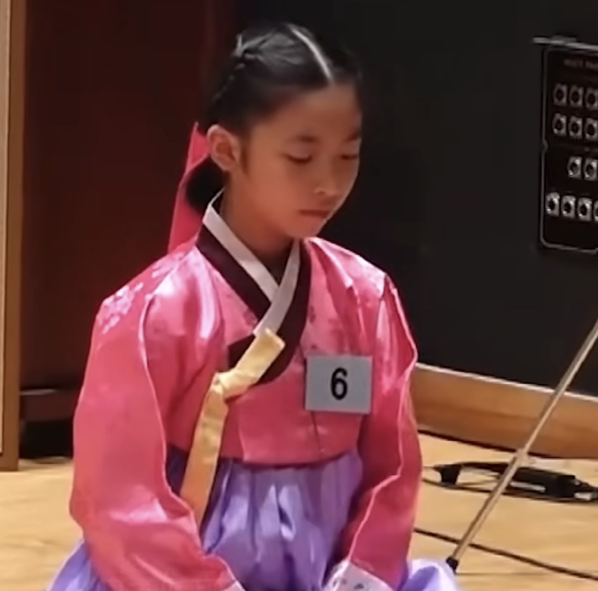 May criança, de hanbok, ajoelhada preparando chá durante a competição