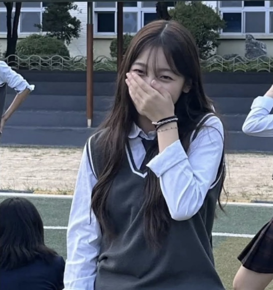 May, de uniforme escolar, rindo com a mão na boca na quadra da escola
