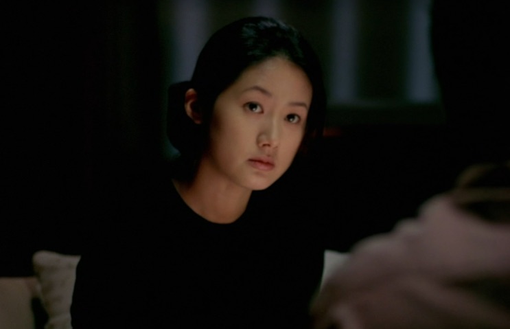 Shim Eun-ha sentada à mesa, olhando para frente, em cena de pouca luz