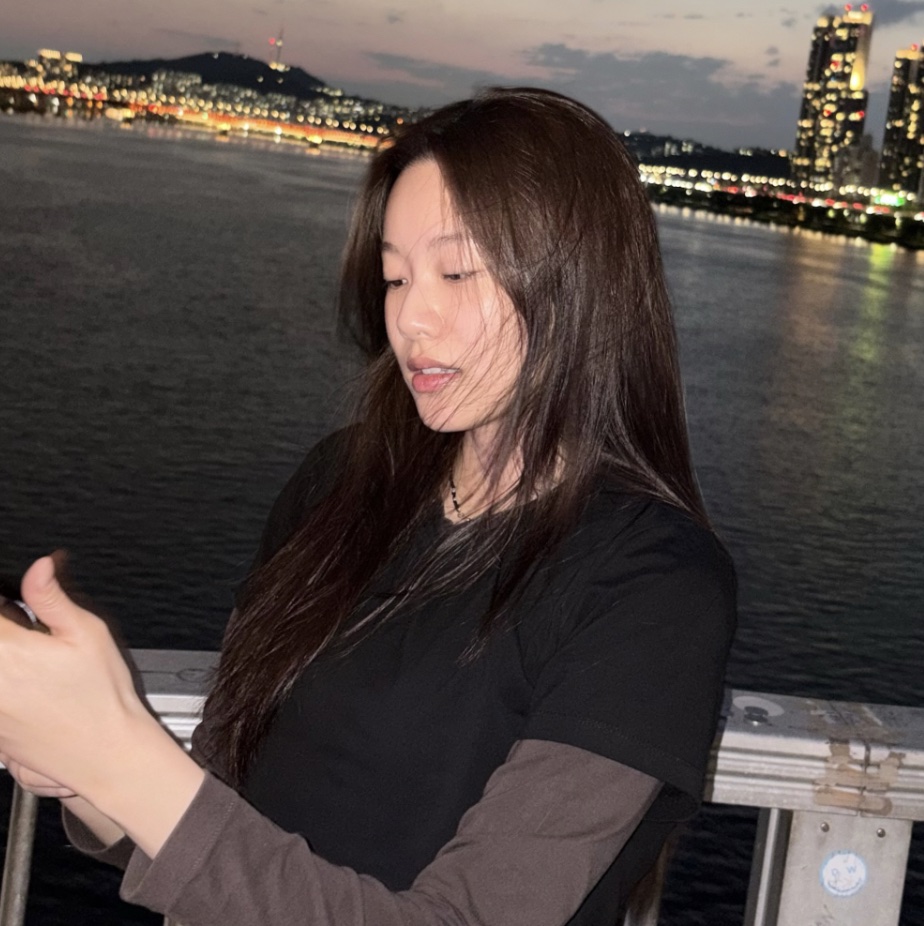 Park Yoo-na à beira de um rio à noite, com prédios iluminados ao fundo