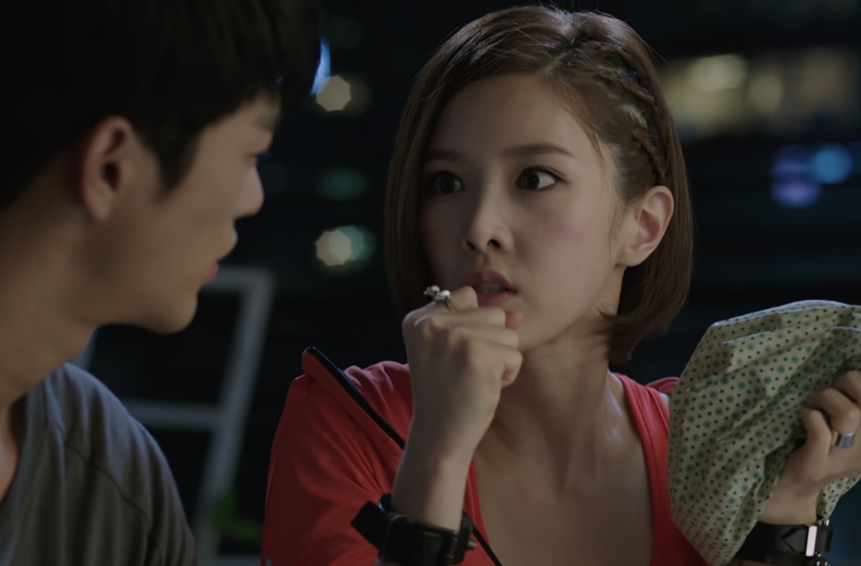 Tae Yi-ryung de blusa vermelha comendo alguma coisa na rua à noite, de frente para Kang Woo, que aparece de costas