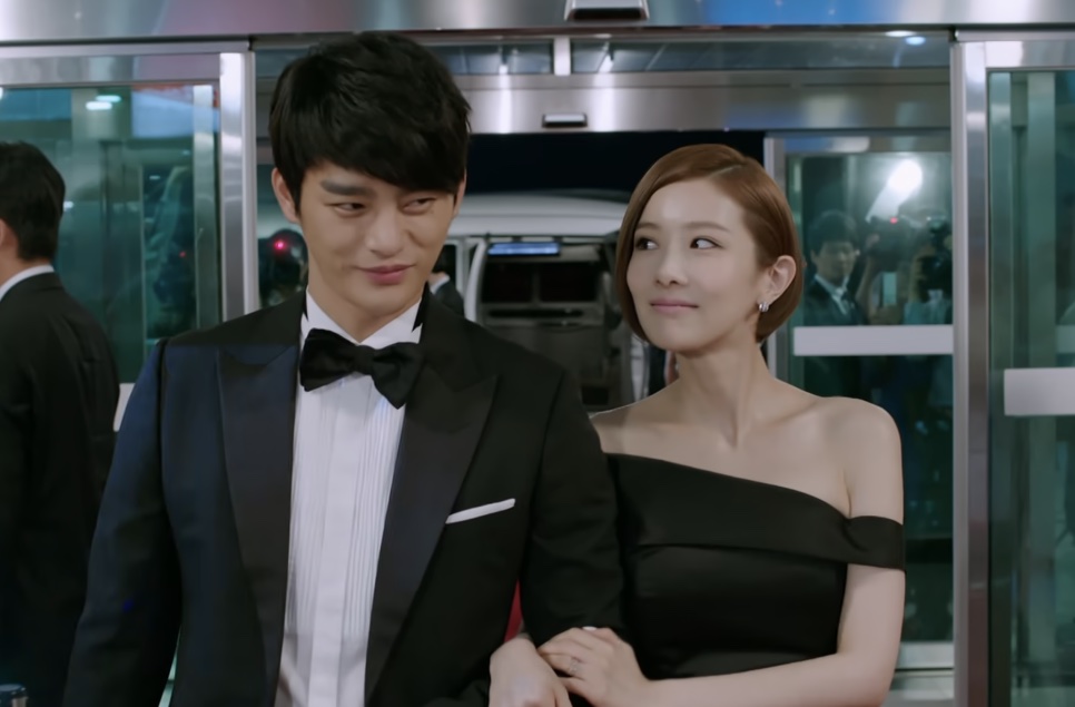 Kang Woo de smoking e Tae Yi-ryung de vestido preto entrando de braços dados, com fotógrafos em volta