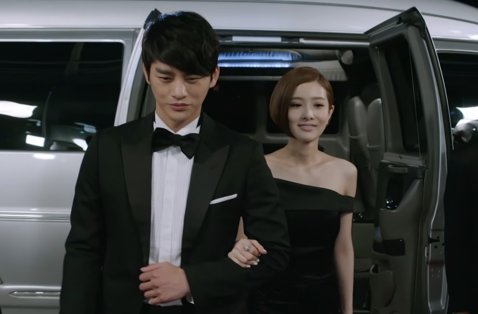 Kang Woo de smoking e Tae Yi-ryung de vestido preto descendo de uma van, de braços dados