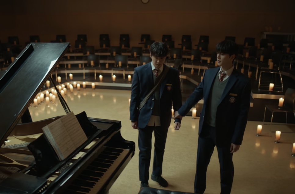 Os dois protagonistas de Quatro Mãos, Duas Sonatas andando num auditório vazio com velas no chão, ao lado de um piano de cauda