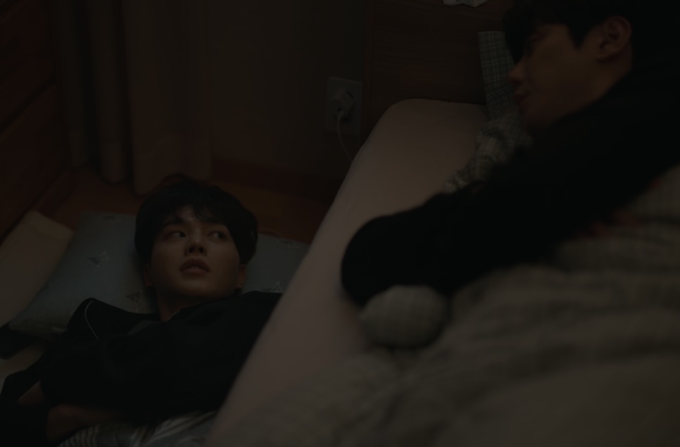 Um dos protagonistas de Quatro Mãos, Duas Sonatas deitado na cama, olhando para o outro, sentado ao lado, num quarto escuro