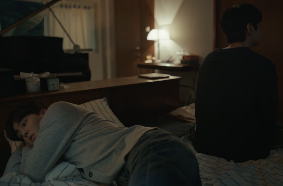 Um dos protagonistas de Quatro Mãos, Duas Sonatas deitado na cama, enquanto o outro está sentado de costas, num quarto com um piano