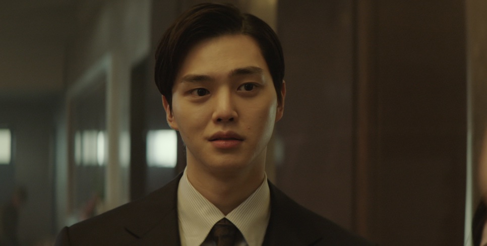 Song Kang de terno e gravata, com o cabelo penteado para trás, no papel de Seon Woo-min no filme Escape