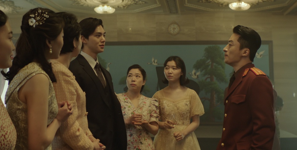 Song Kang de terno no meio de mulheres de vestido, diante de um oficial de uniforme vermelho, numa cena do filme Escape