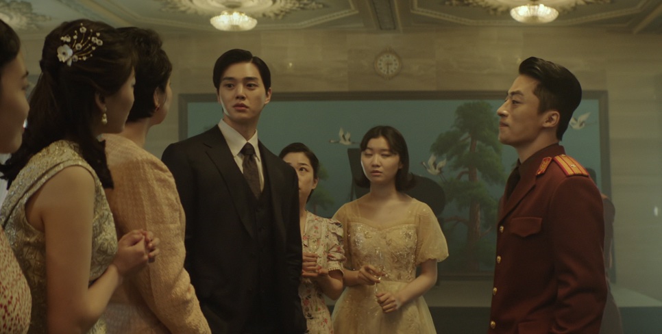 Song Kang de terno escuro num salão, cercado de pessoas bem vestidas, numa cena do filme Escape