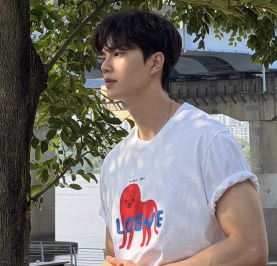 Song Kang de camiseta branca olhando para o lado, debaixo de uma árvore, com um rio ao fundo