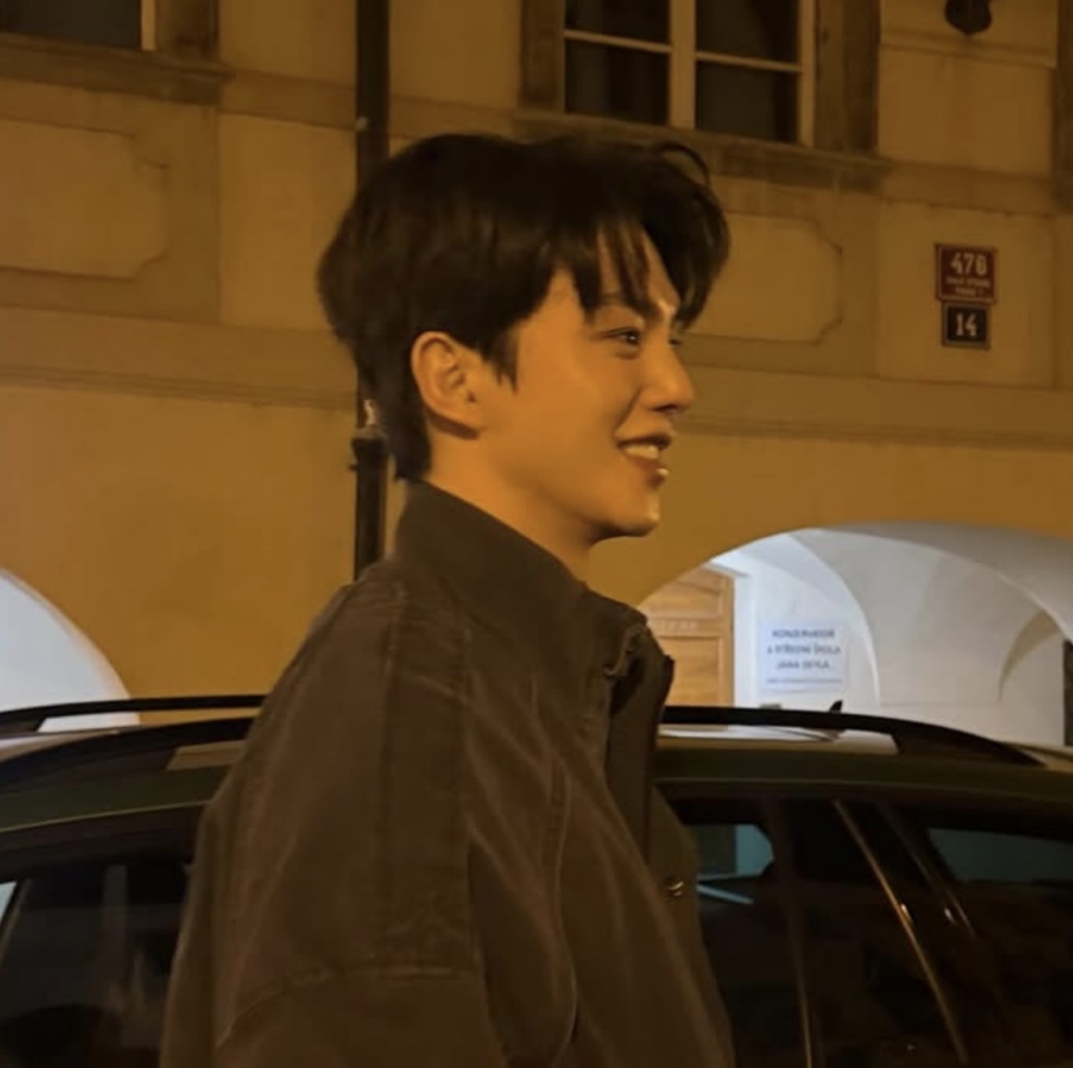 Song Kang sorrindo de perfil, à noite, numa rua com prédios antigos