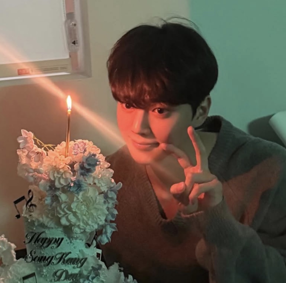Song Kang fazendo sinal de paz ao lado de um bolo de aniversário com uma vela acesa