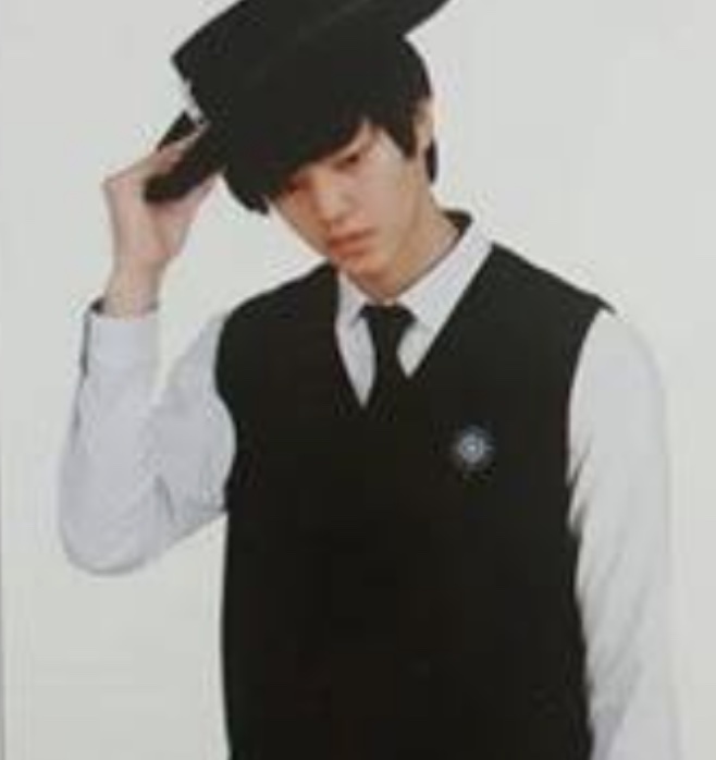 Foto antiga do Song Kang de colete preto e gravata, segurando um chapéu na cabeça