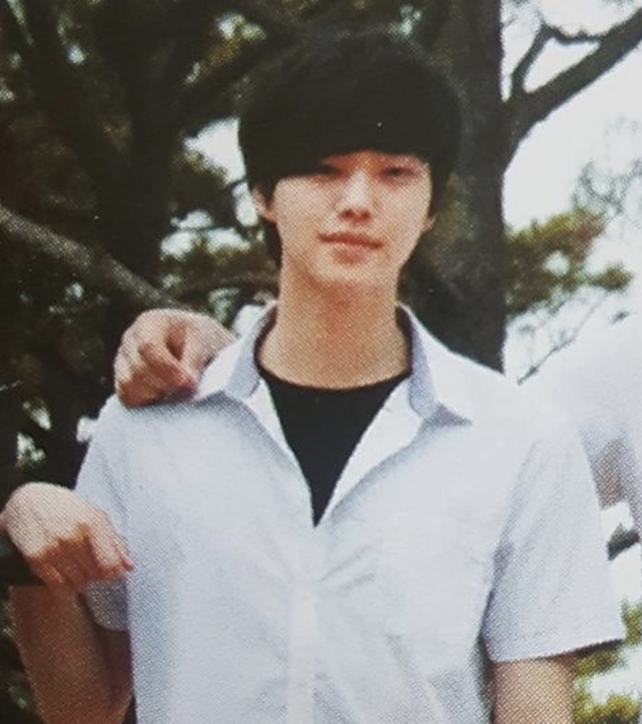 Foto antiga do Song Kang de camisa clara, ao ar livre, com a mão de alguém no ombro dele