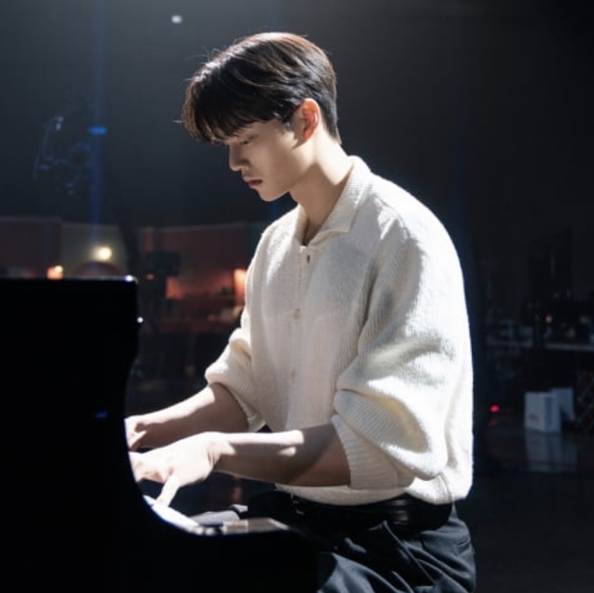 Song Kang de suéter claro tocando piano de cauda num palco escuro, no papel de Kang Pi-o