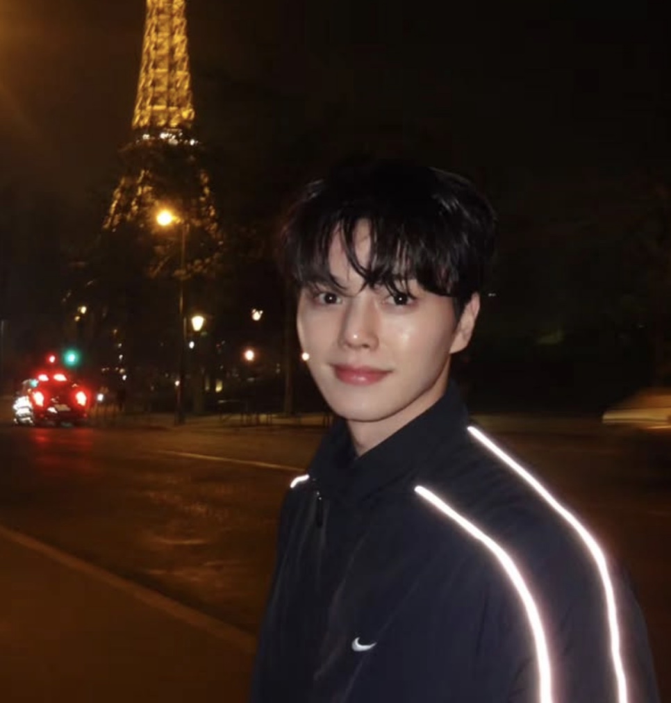 Song Kang de jaqueta escura sorrindo à noite, com uma torre iluminada ao fundo