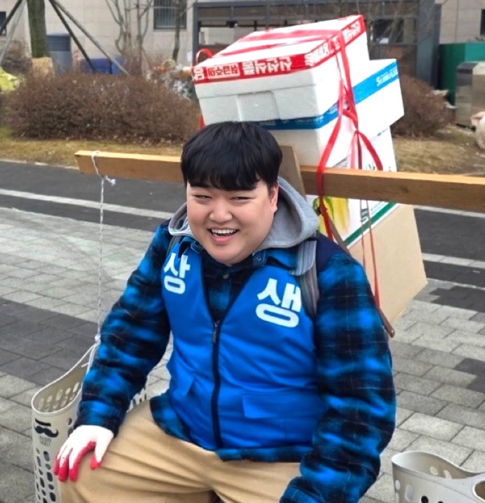 Kim Kyu-won sorrindo, de colete azul, carregando caixas de entrega