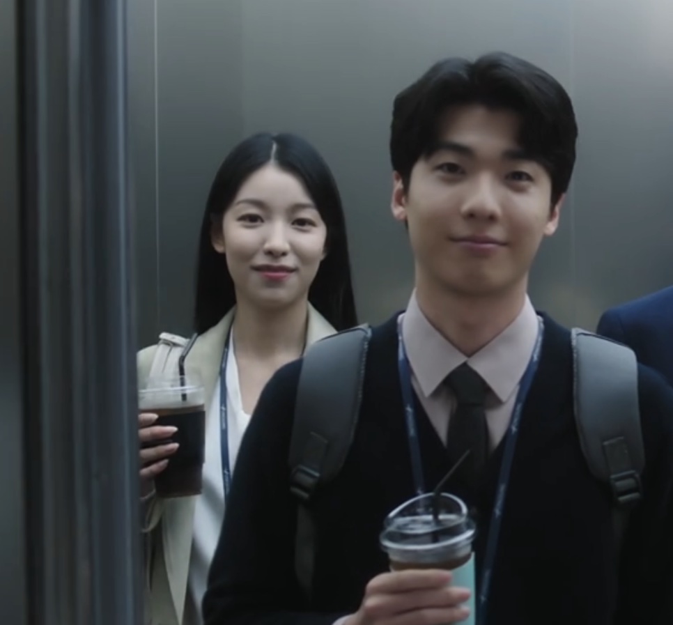 Jang Hyang-eun sorrindo no elevador ao lado de um colega de mochila, os dois segurando copos de café, em The Ordinary Jackpot