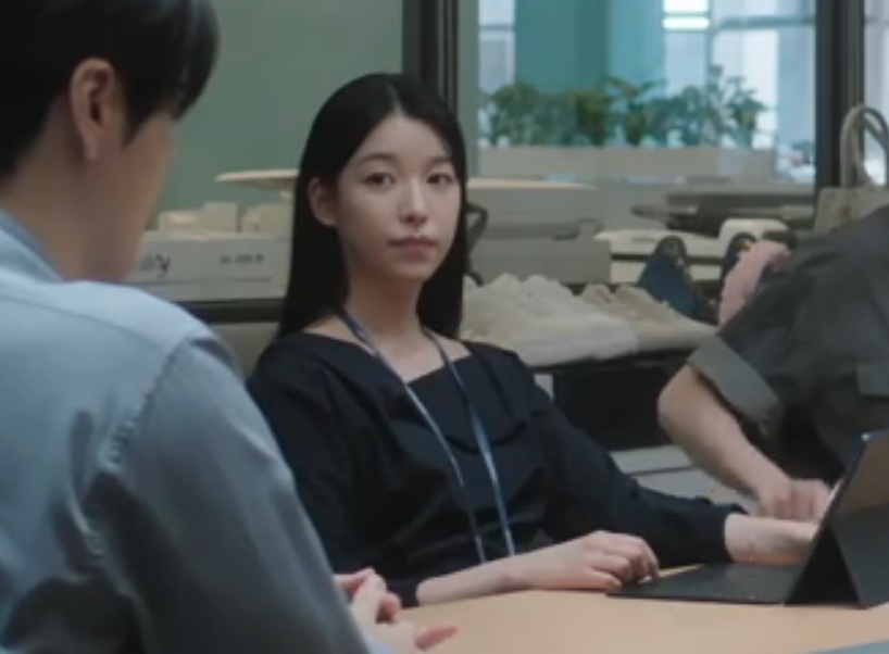 Jang Hyang-eun sentada numa mesa do escritório, de blusa escura e crachá, em The Ordinary Jackpot