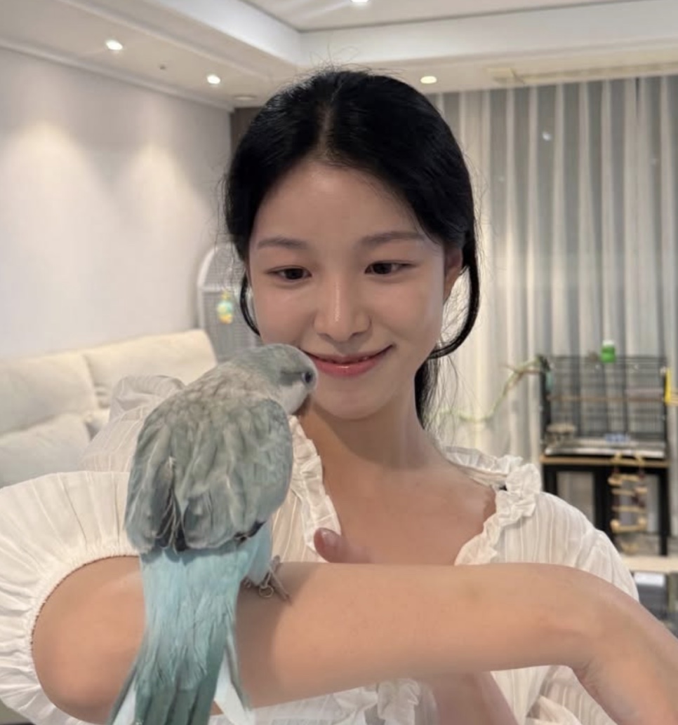 A atriz Baek Yae-in sorrindo em casa, com um periquito azul pousado no braço
