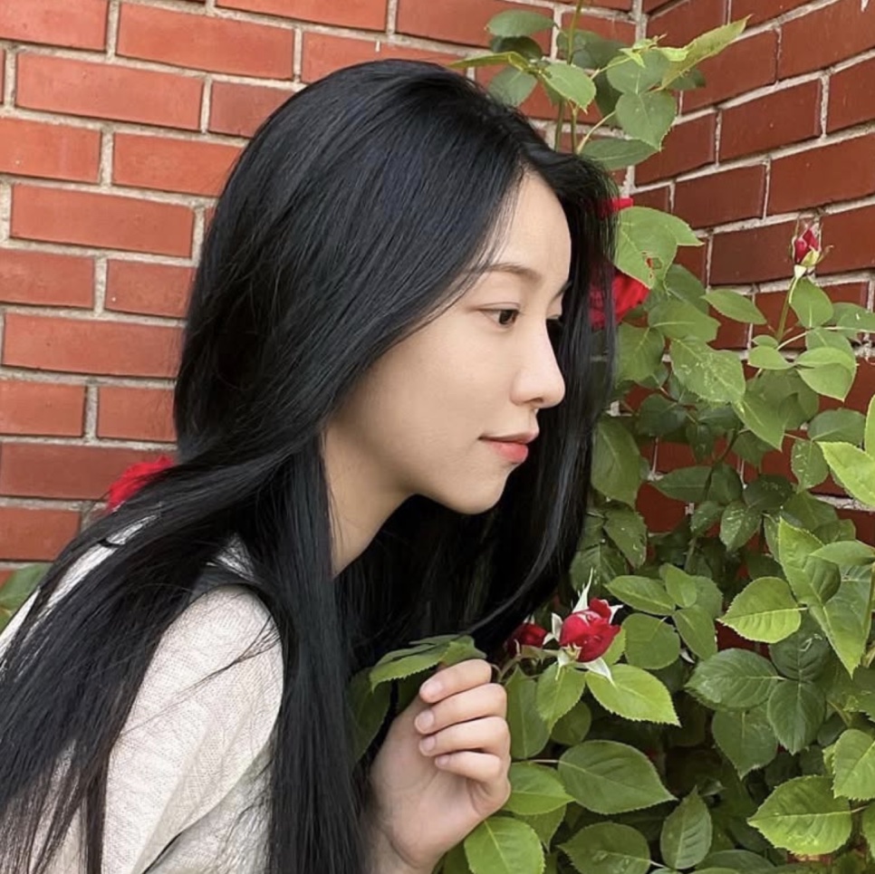 A atriz Baek Yae-in de perfil, cheirando uma rosa vermelha na frente de um muro de tijolos